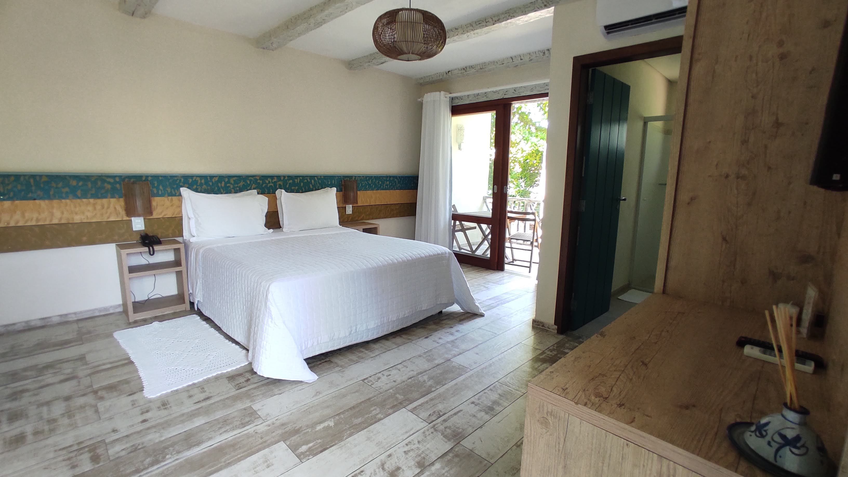 Quarto com cama, mesas de cabeceira, piso de madeira, uma pequena varanda e porta que dá para o exterior.