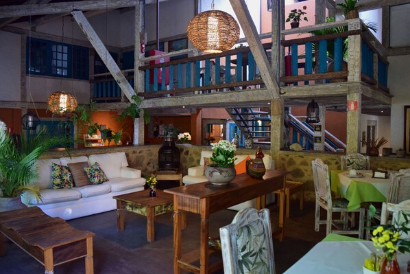Un interior amplio con un techo alto, vigas de madera y una escalera que conduce a un nivel superior. Hay varias zonas de estar con mesas y sillas, y plantas decoran el espacio.