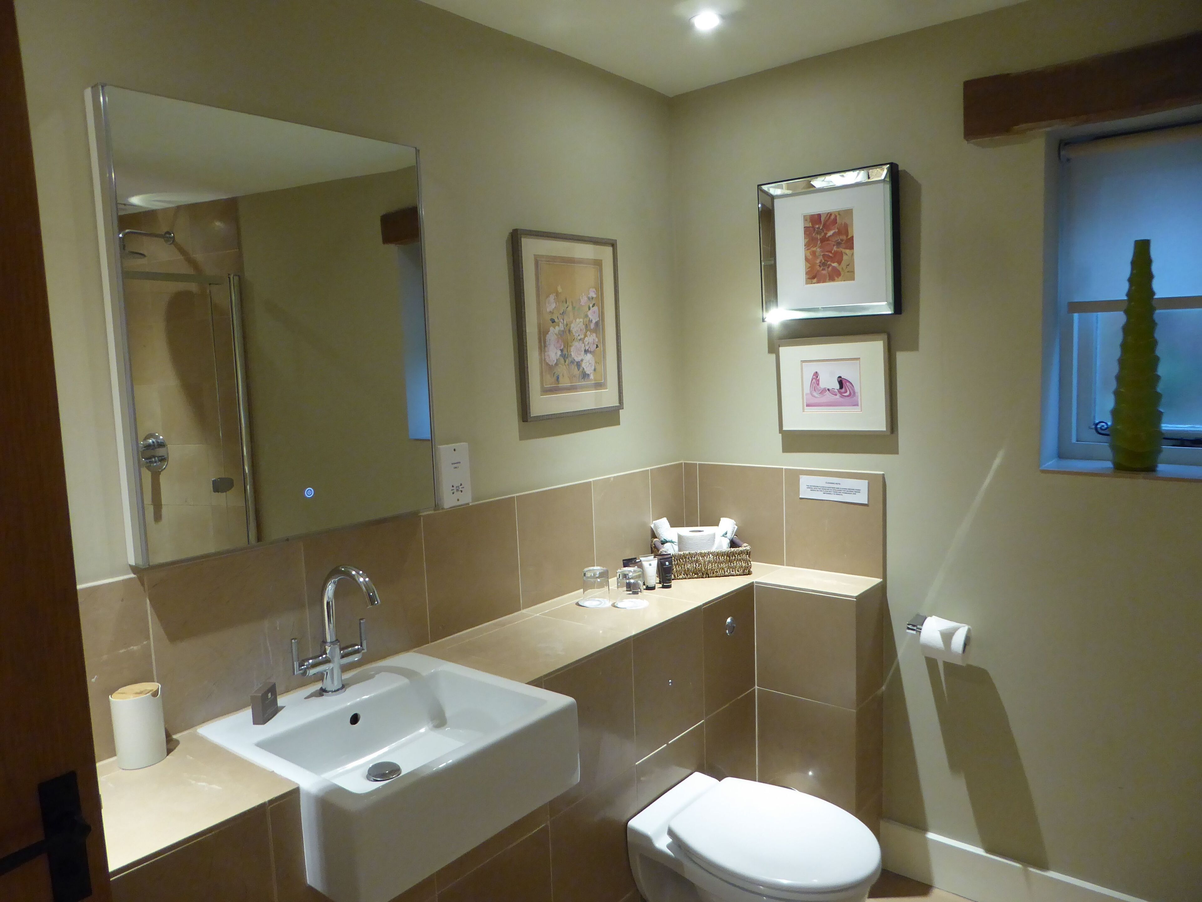 Un baño moderno con lavamanos, inodoro y ducha. Hay cuadros en la pared y una ventana con una planta.