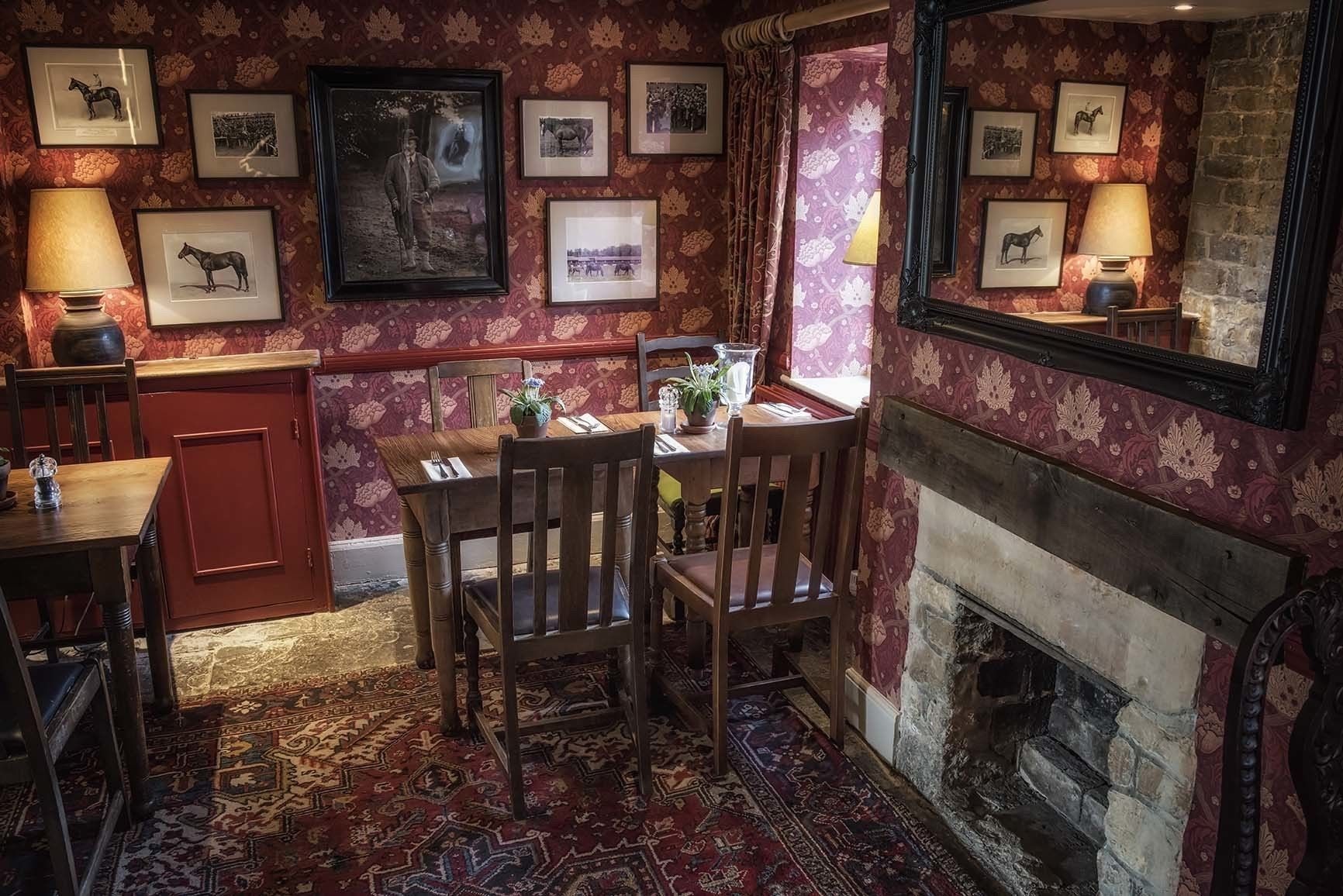 A traditional dining room with a fireplace, patterned wallpaper, and framed equestrian photographs on the walls.