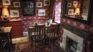 A traditional dining room with a fireplace, patterned wallpaper, and framed equestrian photographs on the walls.