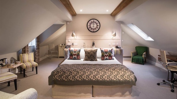 A bedroom with a large bed, a wall clock, a desk with a chair, and a window. - The Howard Arms (Shipston on Stour)