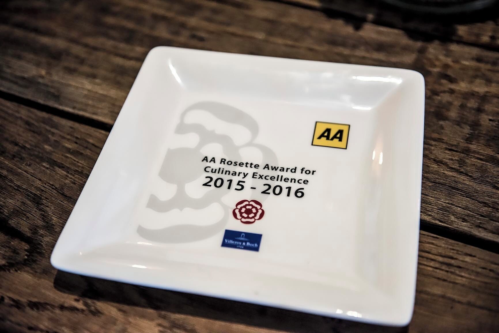 A square white plate with text and logos, placed on a wooden surface.