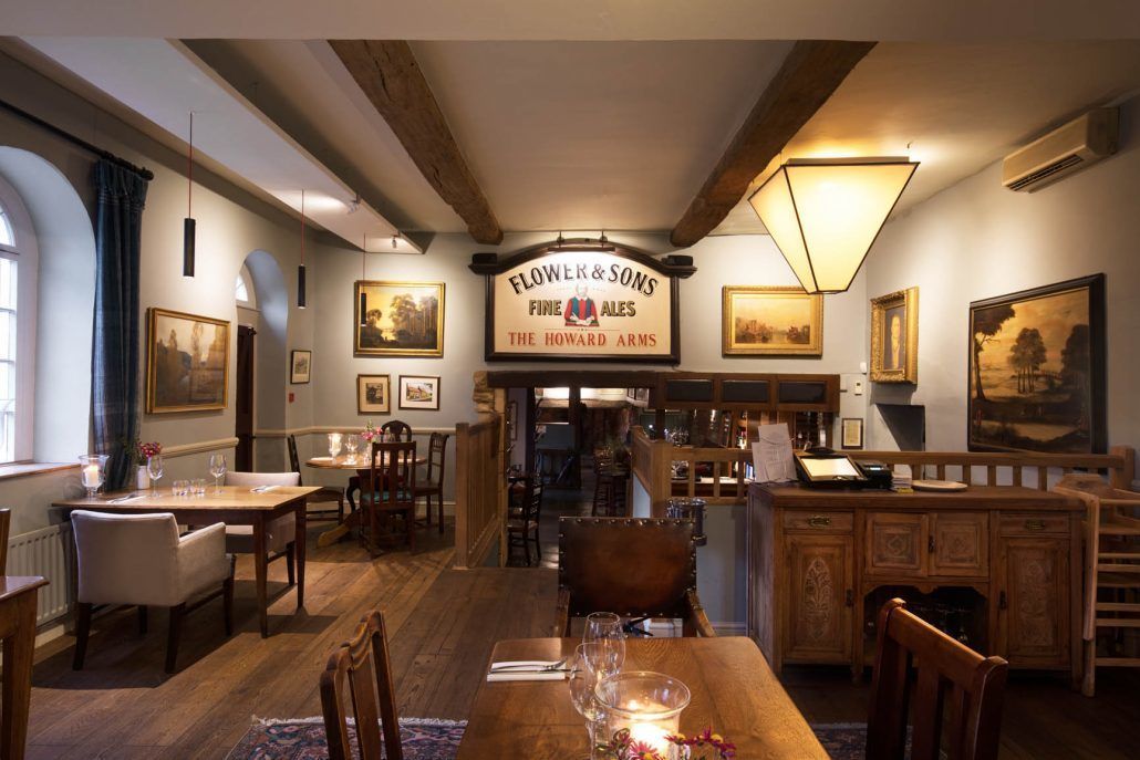 A restaurant interior with wooden tables and chairs, framed artwork on the walls, and a sign reading "Flower & Sons Fine Ales The Howard Arms".