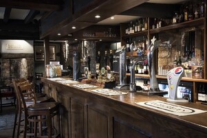 Un área de bar con taburetes de madera, una variedad de botellas y vasos en estantes, y un dispensador de Pepsi.