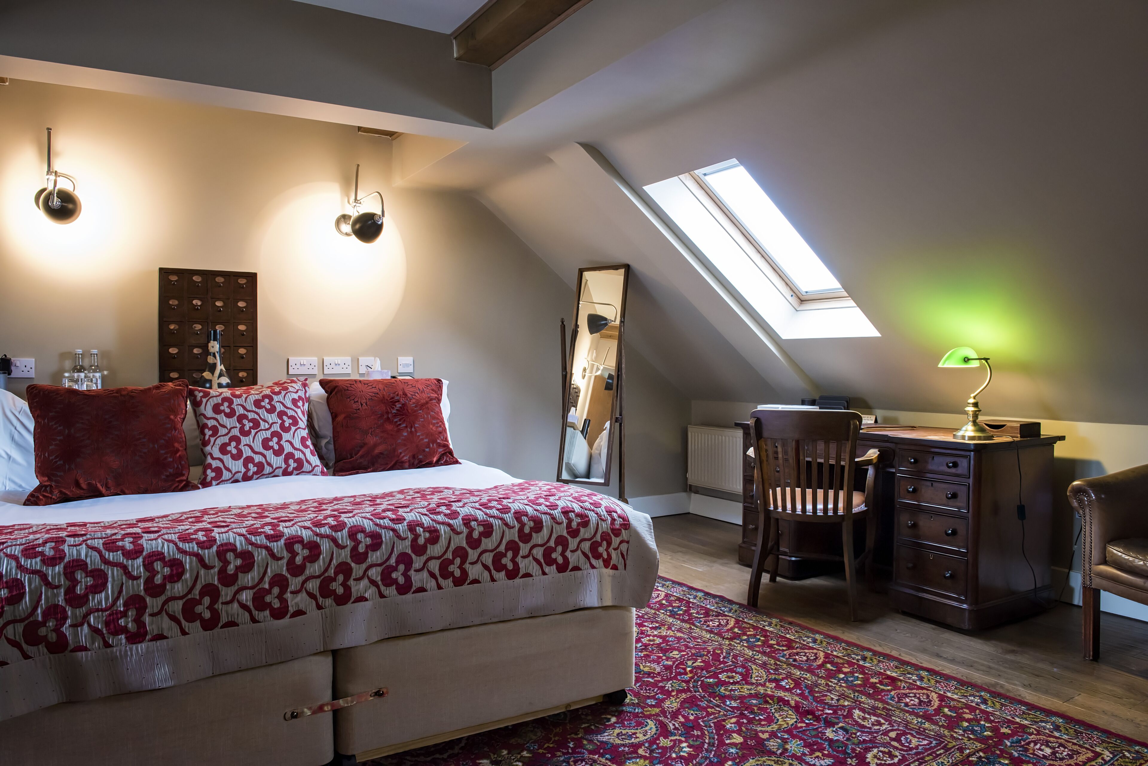 A bedroom with a large bed, a desk, and a chair. There is a skylight above the bed and a mirror on the wall.