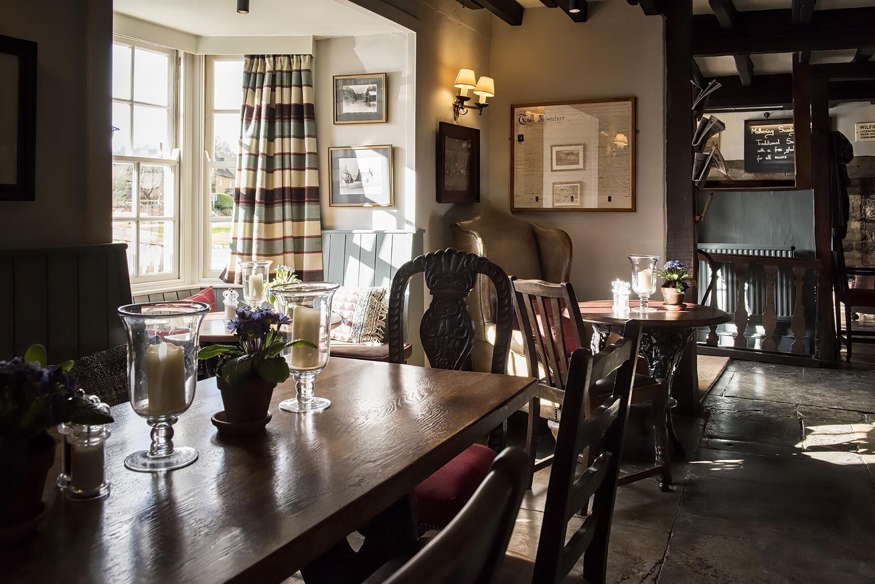 A cozy dining area with a wooden table, chairs, and a window with curtains. There are framed pictures on the wall and a menu board.