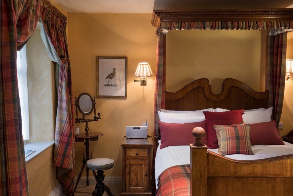 Una habitación tradicional con cama con dosel, una mesita de noche con lámpara, un espejo y una ventana con cortinas.