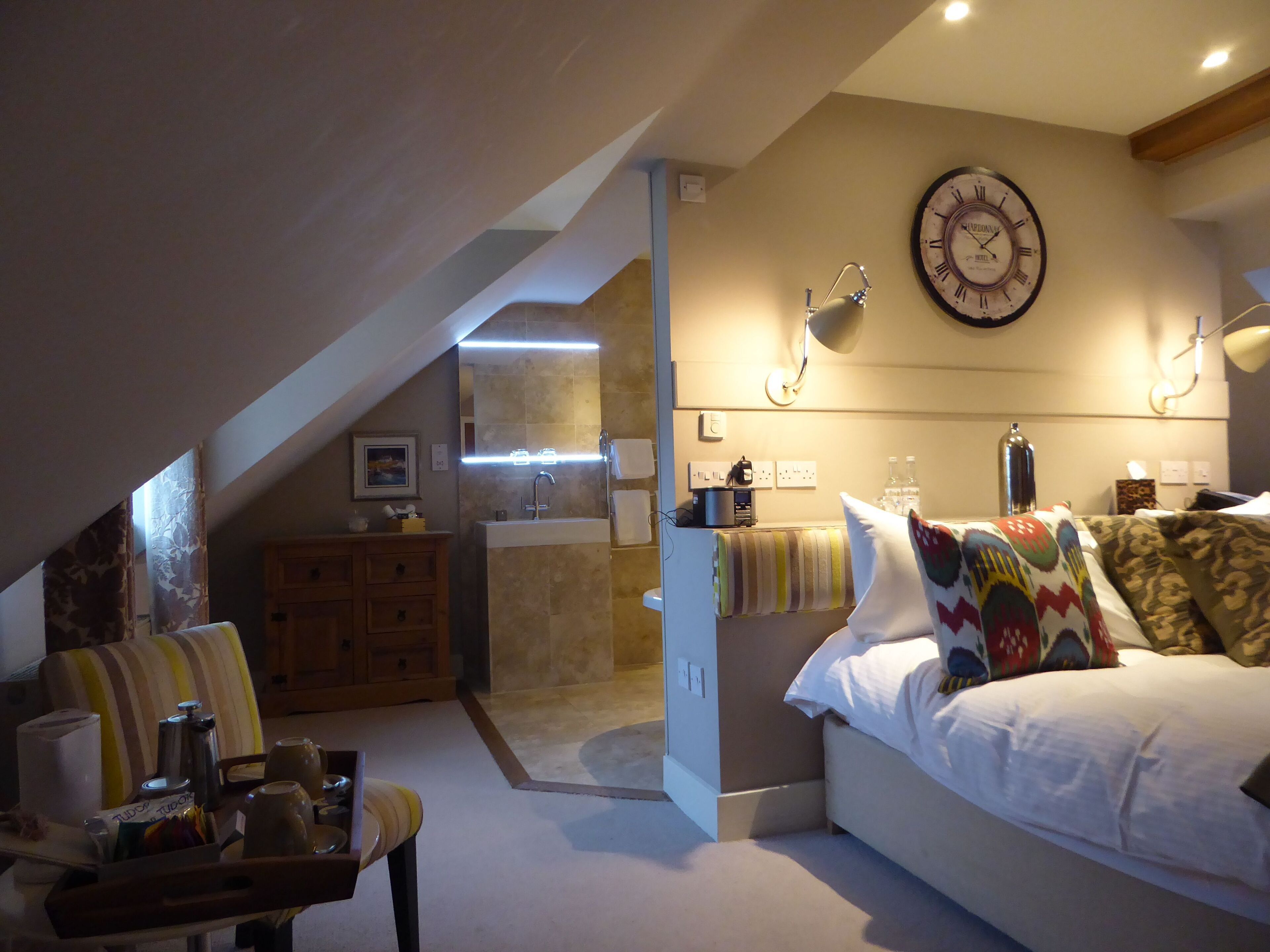 A bedroom with a bed, a nightstand, a lamp, a clock on the wall, and a view of the bathroom.