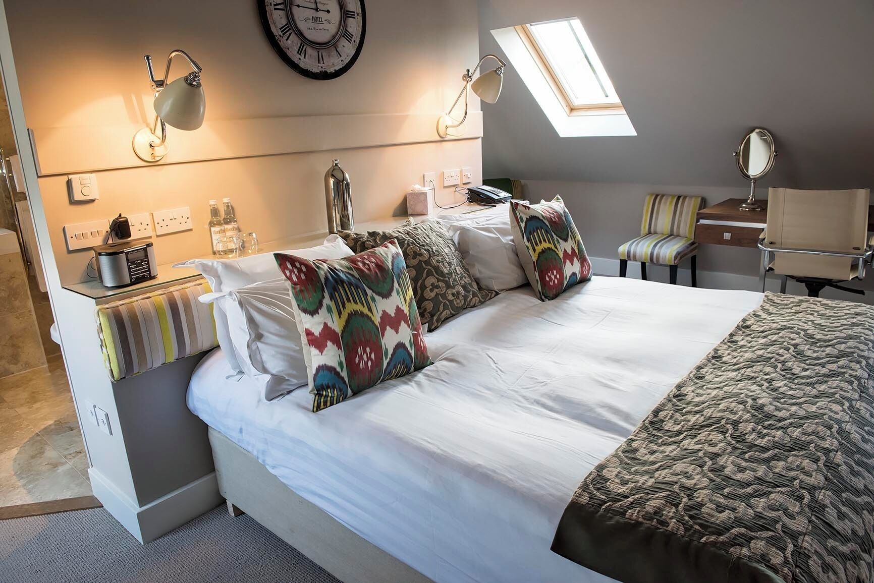 A modern bedroom with a large bed, a clock on the wall, a skylight, and a desk with a lamp.