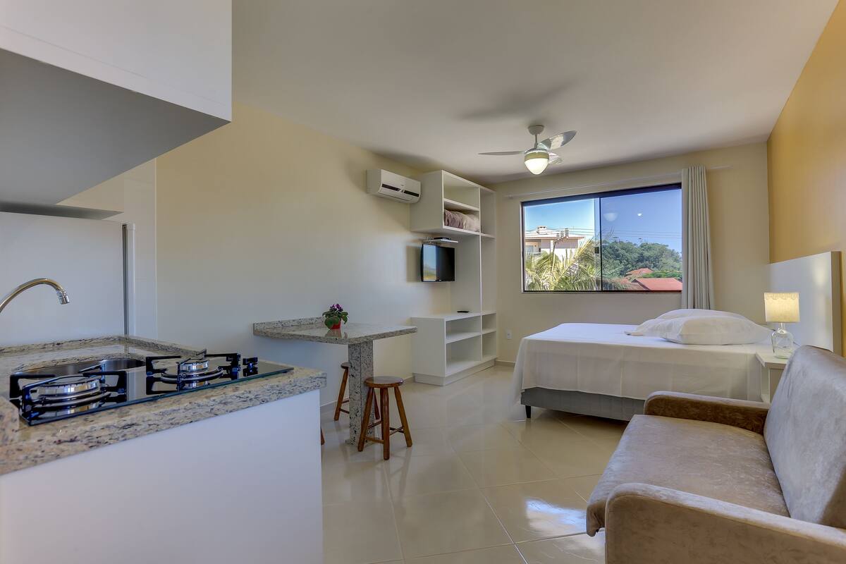 A compact hotel room with a kitchenette, a bed, a sofa, and a large window with a view.