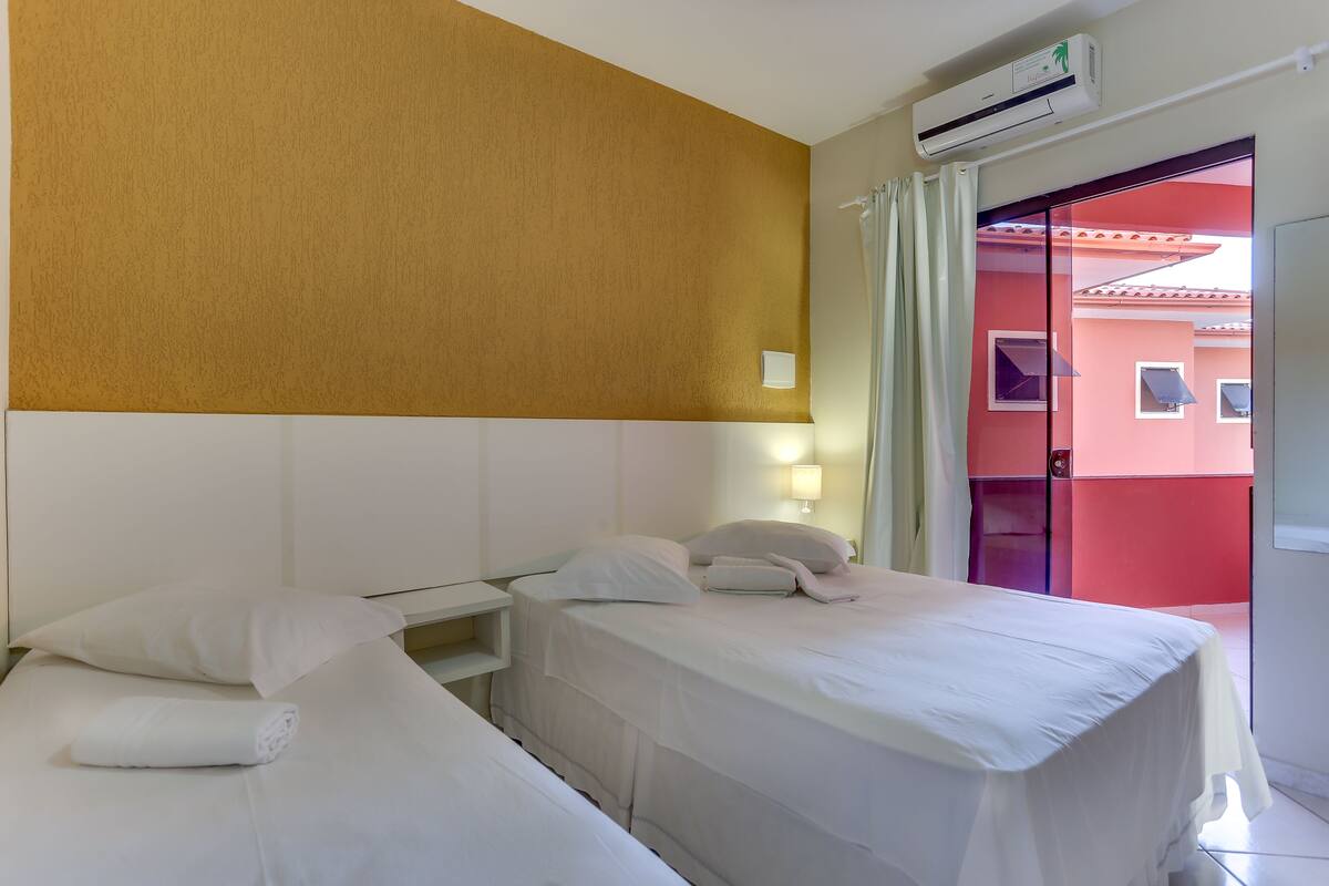 A hotel room with two beds, a white headboard, a wall-mounted air conditioner, and a view of a red building through an open door.