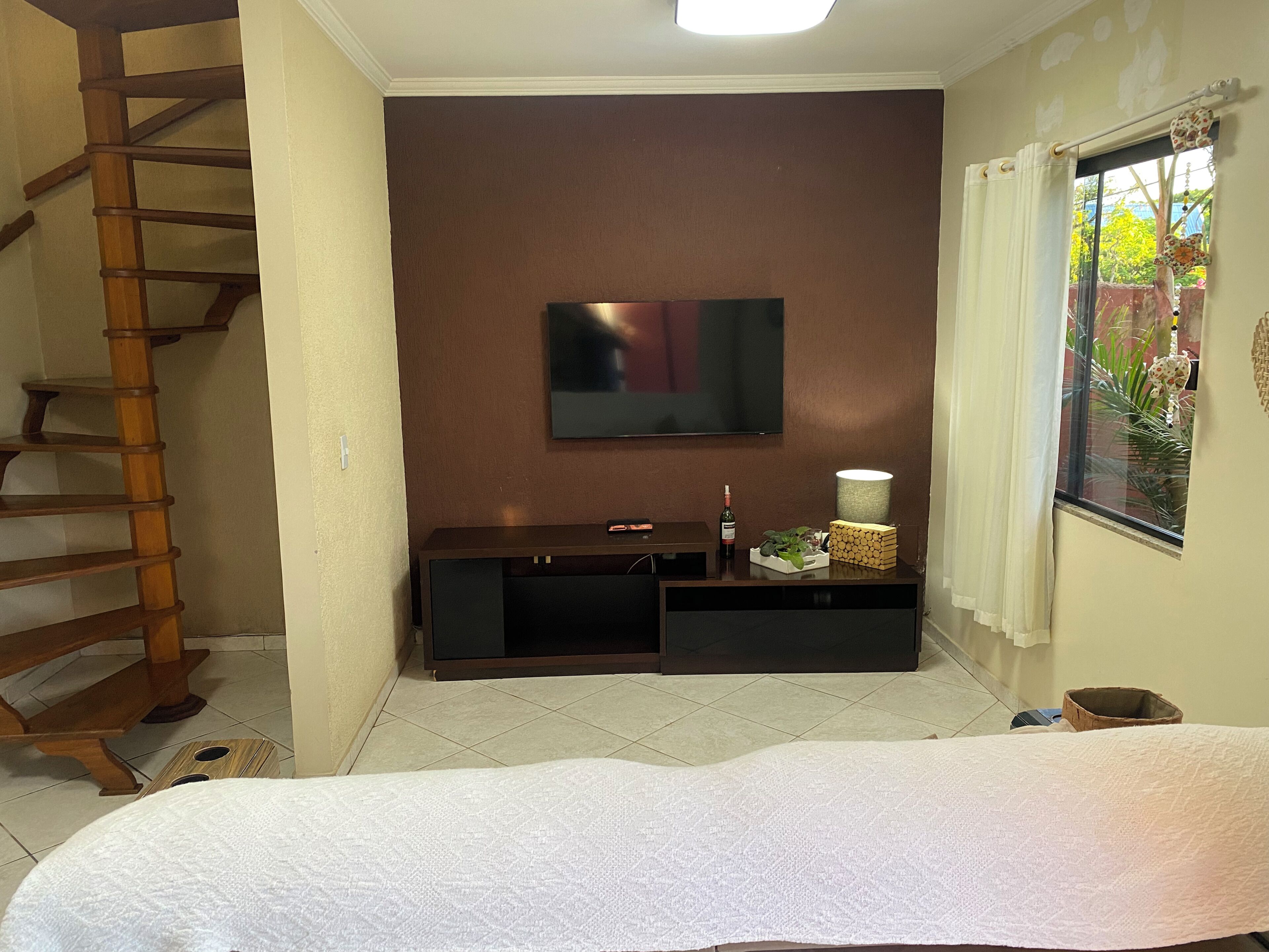 A bedroom with a bed, a TV mounted on the wall, a wooden ladder, a window with curtains, and a small table with a lamp and a bottle.