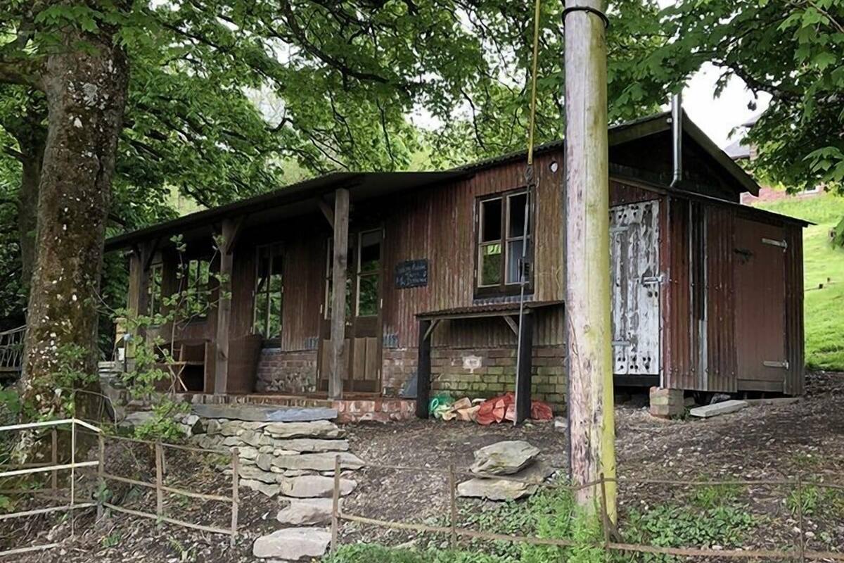 A rustic wooden cabin with a stone staircase leading to a porch, surrounded by trees and a grassy area.