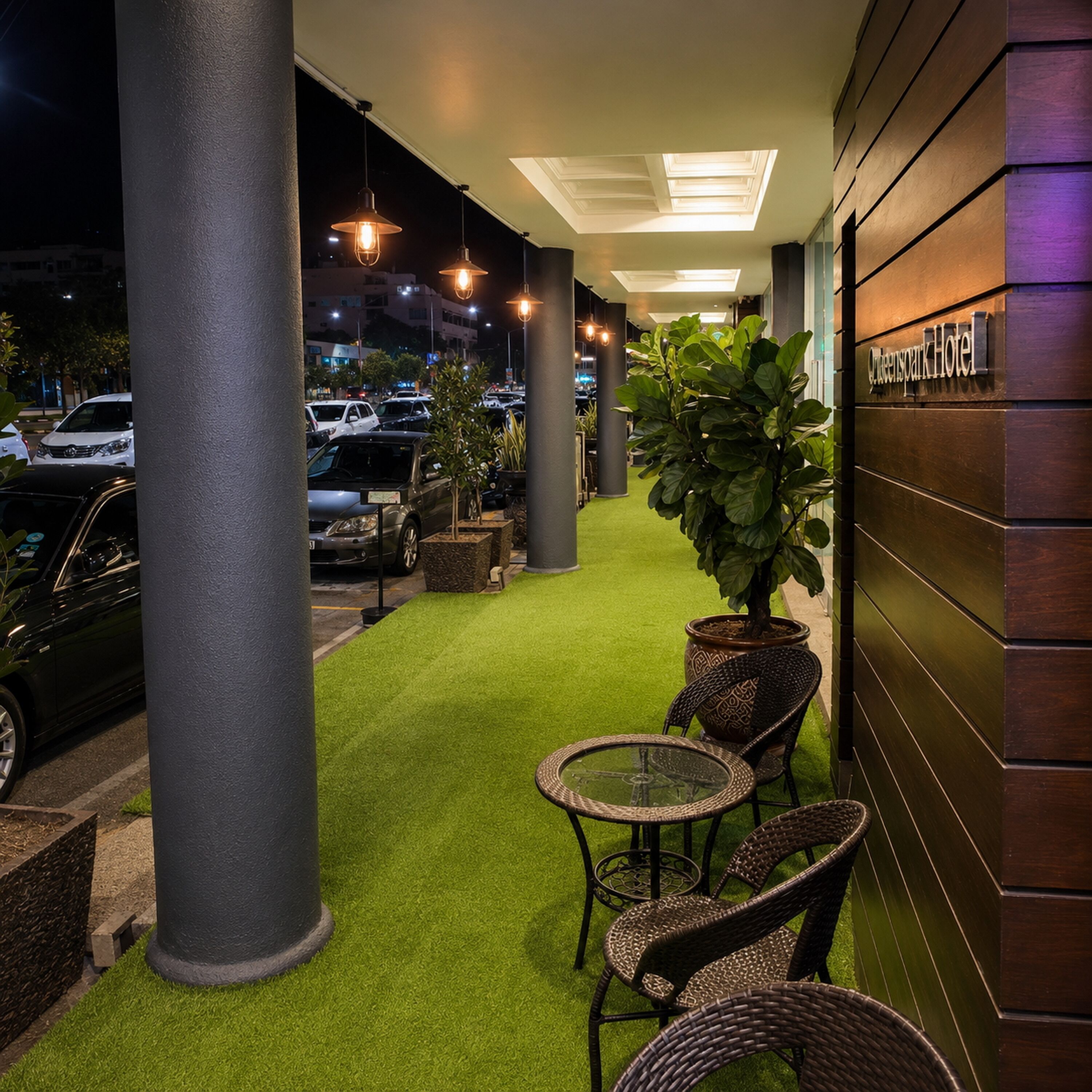 An outdoor seating area with artificial grass, wicker furniture, and a building with a sign reading 'Queenspark Hotel.'