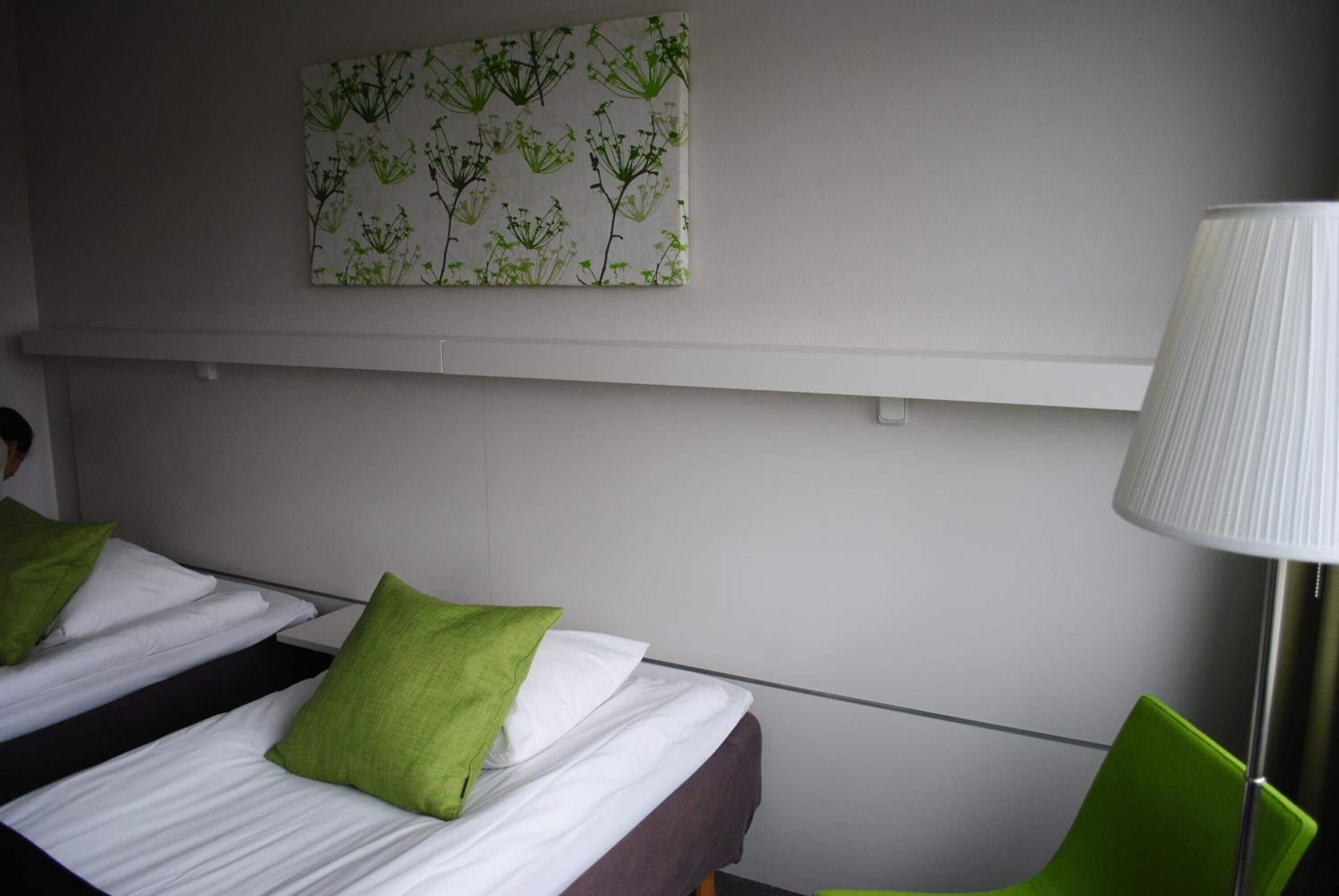 Habitación de hotel con dos camas individuales, cada una con sábanas blancas y almohadas verdes. Hay una silla verde y una lámpara de pie. Se observa una obra de arte en la pared con motivos florales en tonos verdes y blancos sobre las camas.