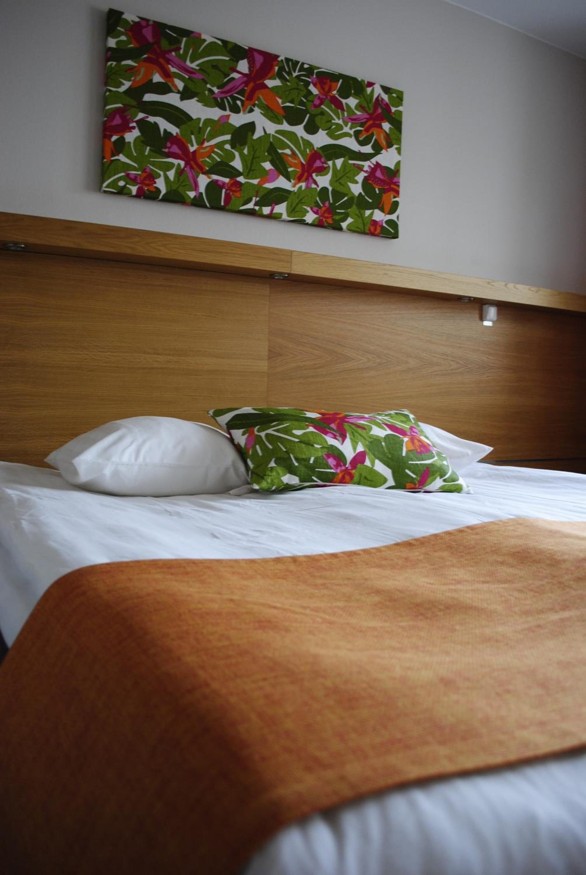 A hotel room with a wooden headboard, a bed with white bedding, a colorful floral pillow, and a large floral artwork on the wall.