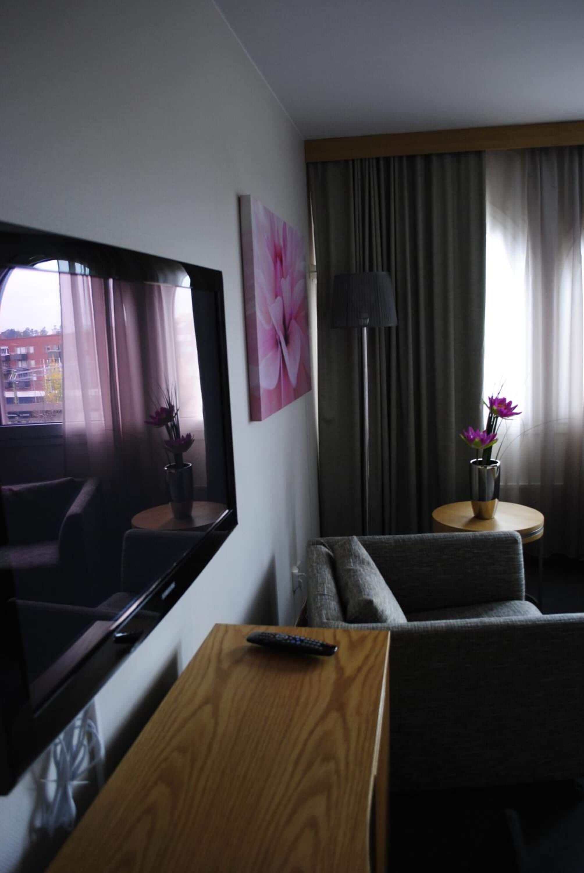 Hotelkamer met televisie, bank, klein tafeltje, afstandsbediening en een vaas met bloemen.