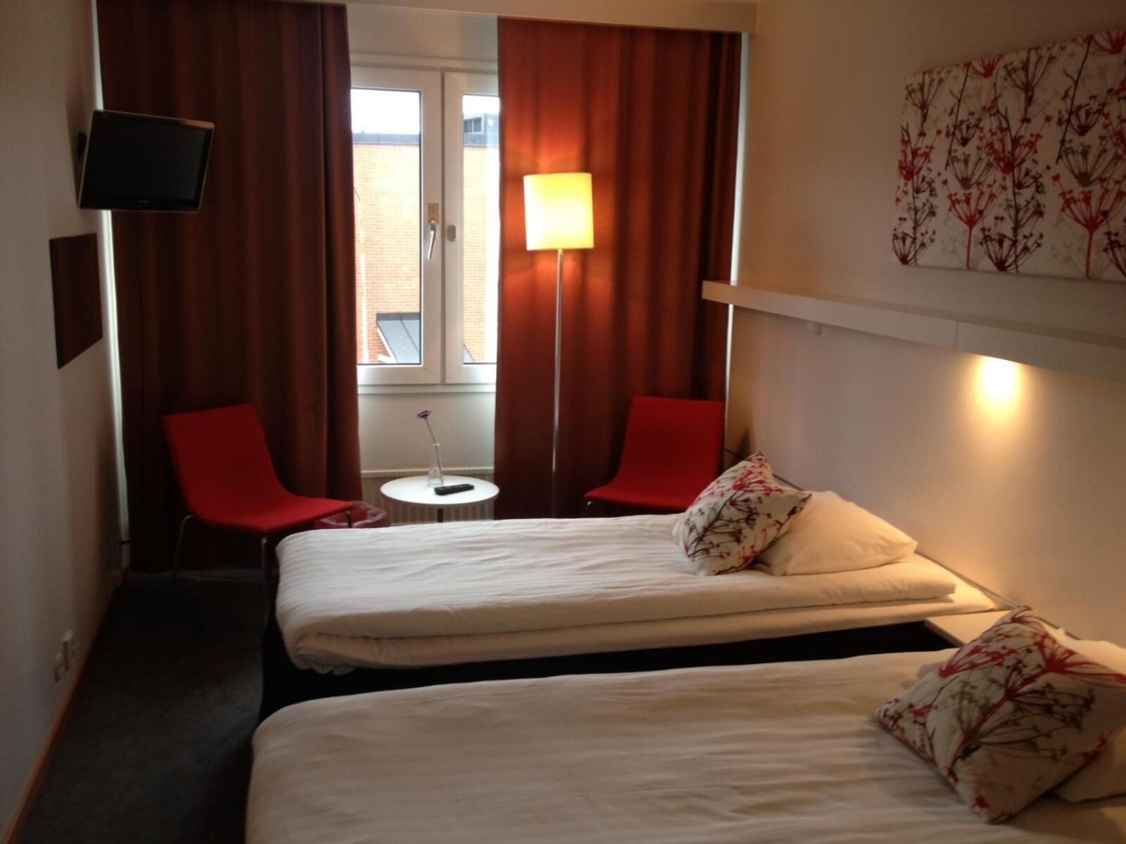 A hotel room with two beds, a TV, a small table, and two red chairs.