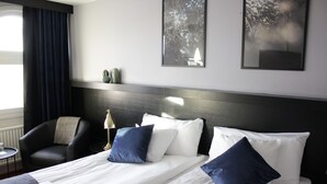 Habitación de hotel con cama, dos almohadas, una silla, una mesita y dos cuadros colgados en la pared.