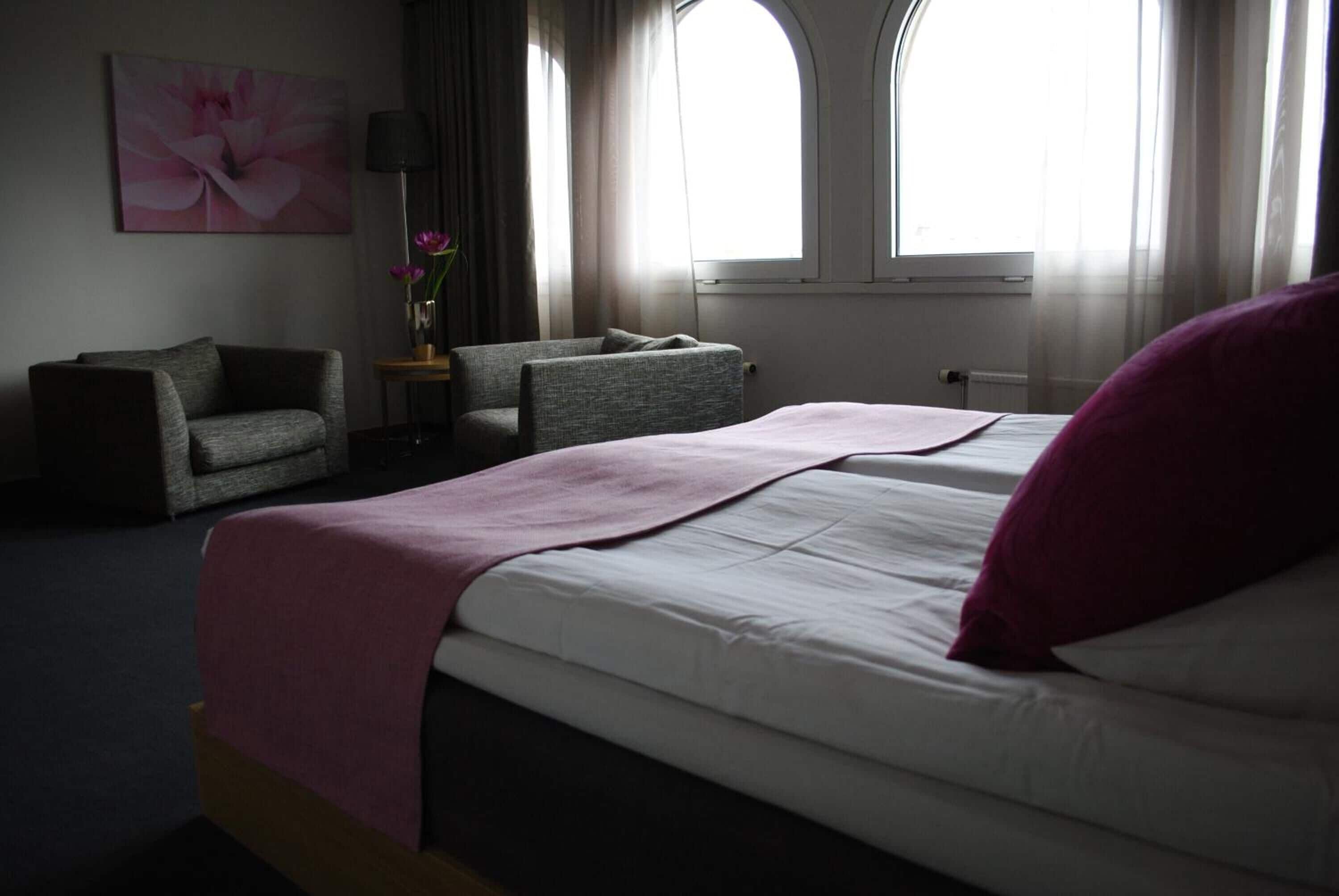 A hotel room with a bed, a chair, a small table with a vase of flowers, and two windows with curtains.