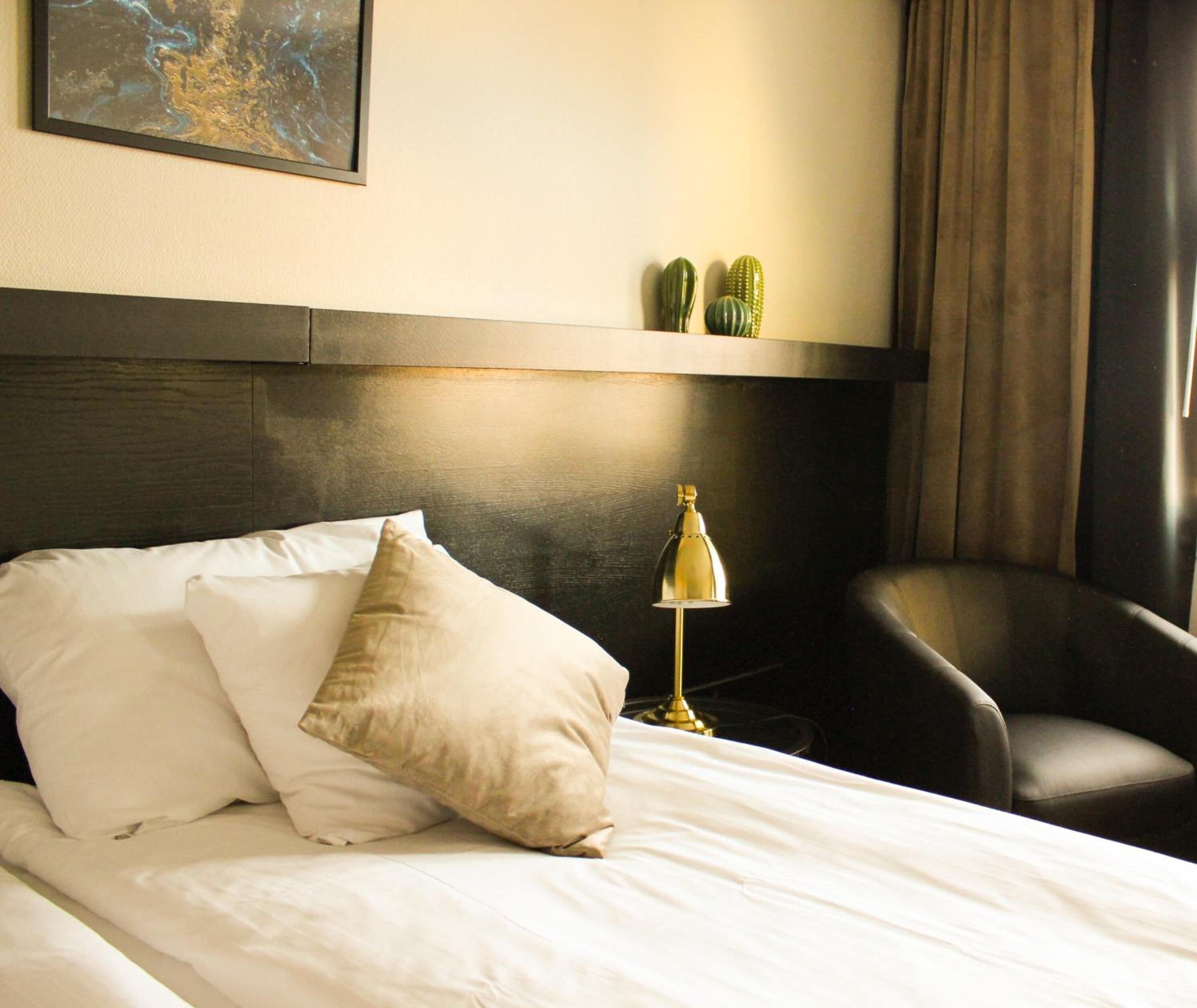 Camera d'albergo con un letto, una scrivania con lampada, una sedia e una mensola a parete con oggetti decorativi.