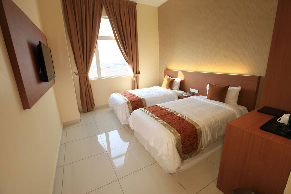 A hotel room with two beds, a desk, a TV mounted on the wall, and a window with curtains.