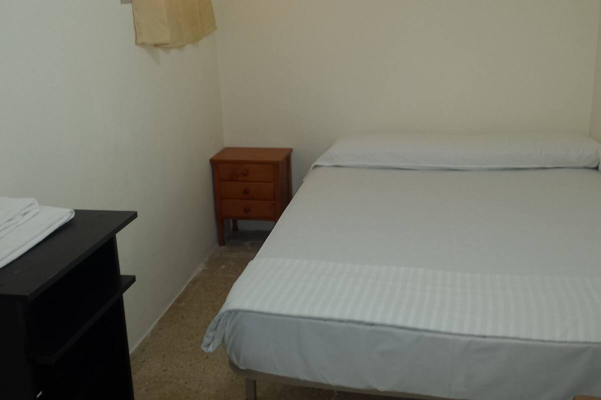 Habitació petita i poc moblada, amb un llit, una tauleta de nit i un escriptori amb una tovallola blanca plegada.