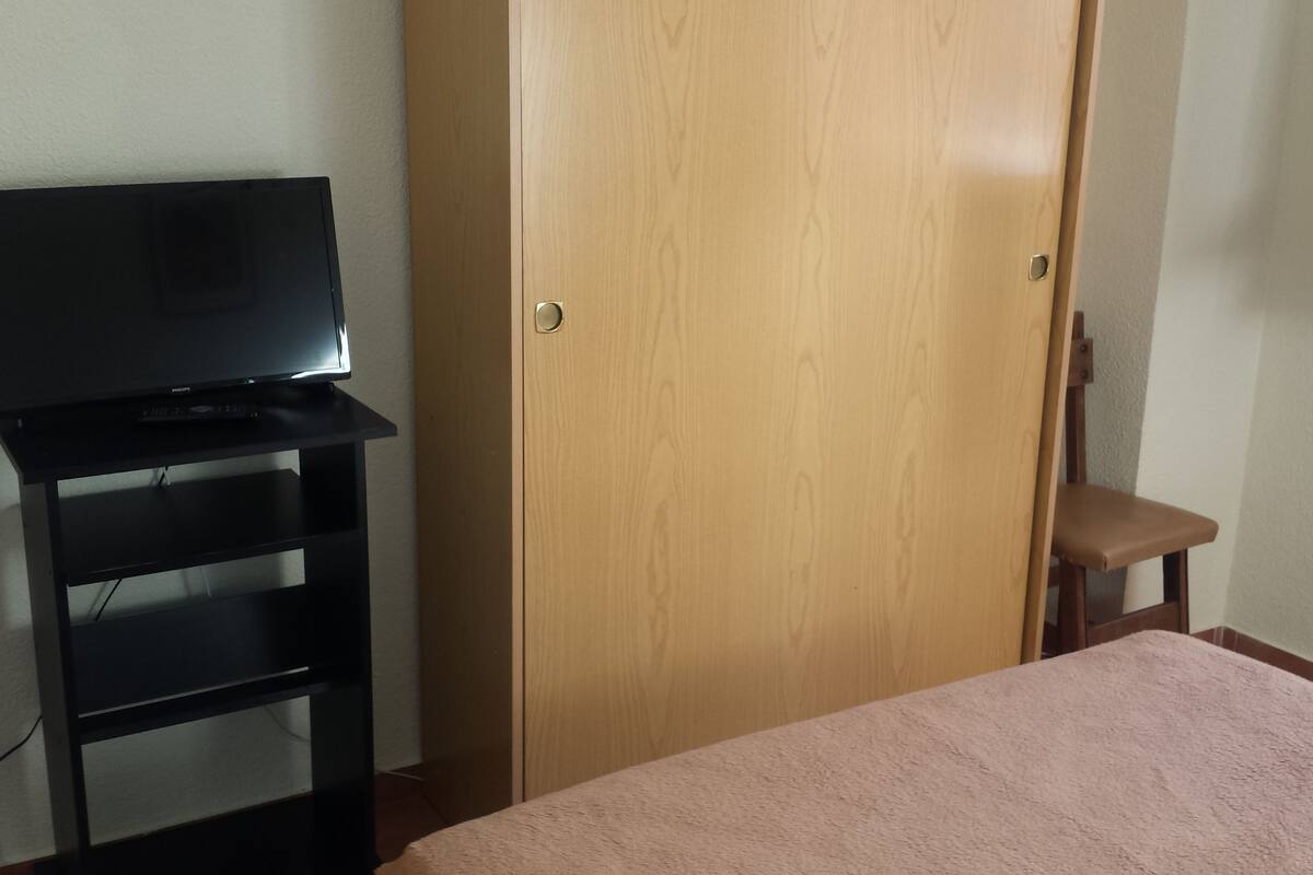 Una habitació amb un llit, un armari de fusta, un prestatge negre per a la televisió i una cadira.