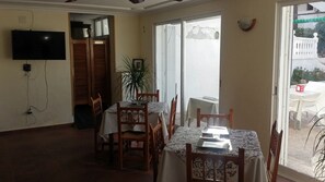 Zona pranzo con tavolo apparecchiato per quattro, sedie di legno, una pianta in vaso e un televisore montato a parete.
