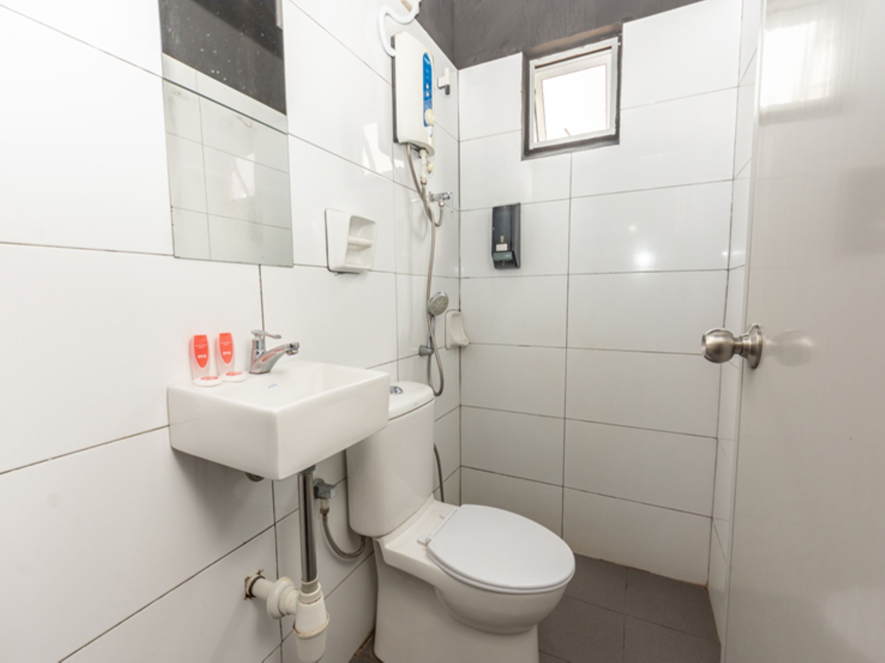 Un baño con lavamanos, inodoro y ducha de color blanco. Hay una ventana pequeña sobre el inodoro.