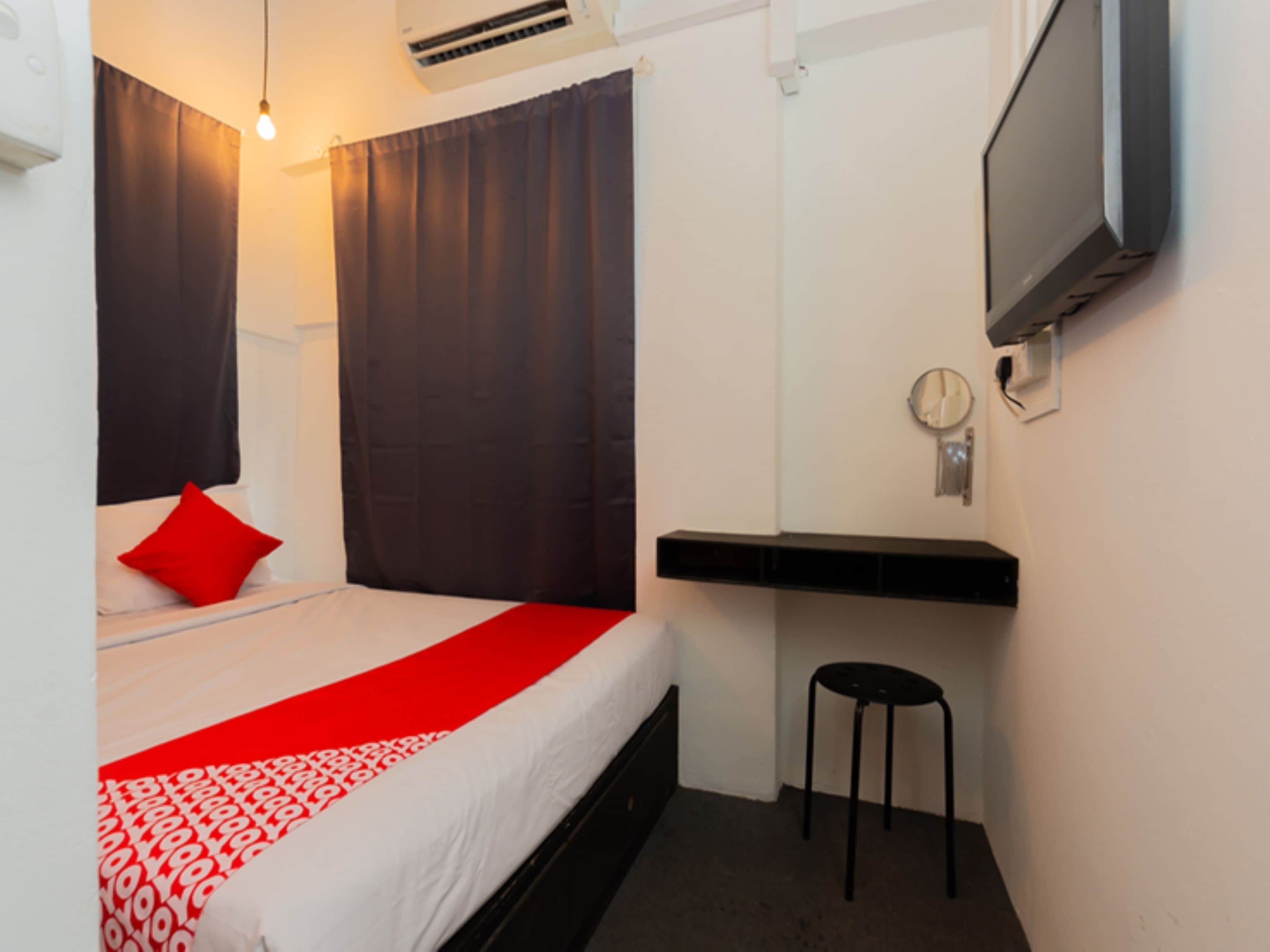 A hotel room with a bed, a red and white patterned bedspread, a black and white wall, a mounted television, and a small stool.