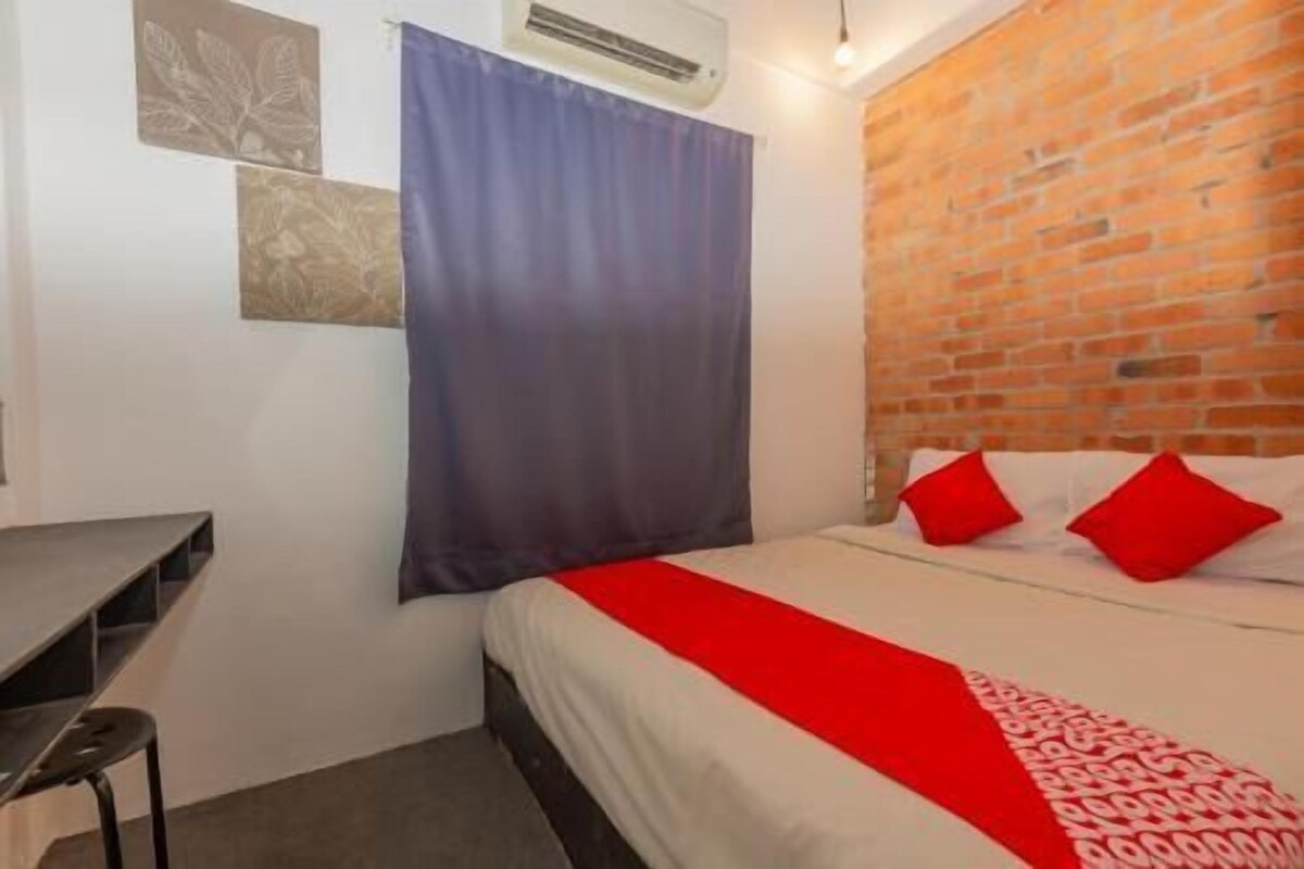 Un dormitorio con pared de ladrillo, cama con almohadas rojas, colcha blanca y cortina azul.