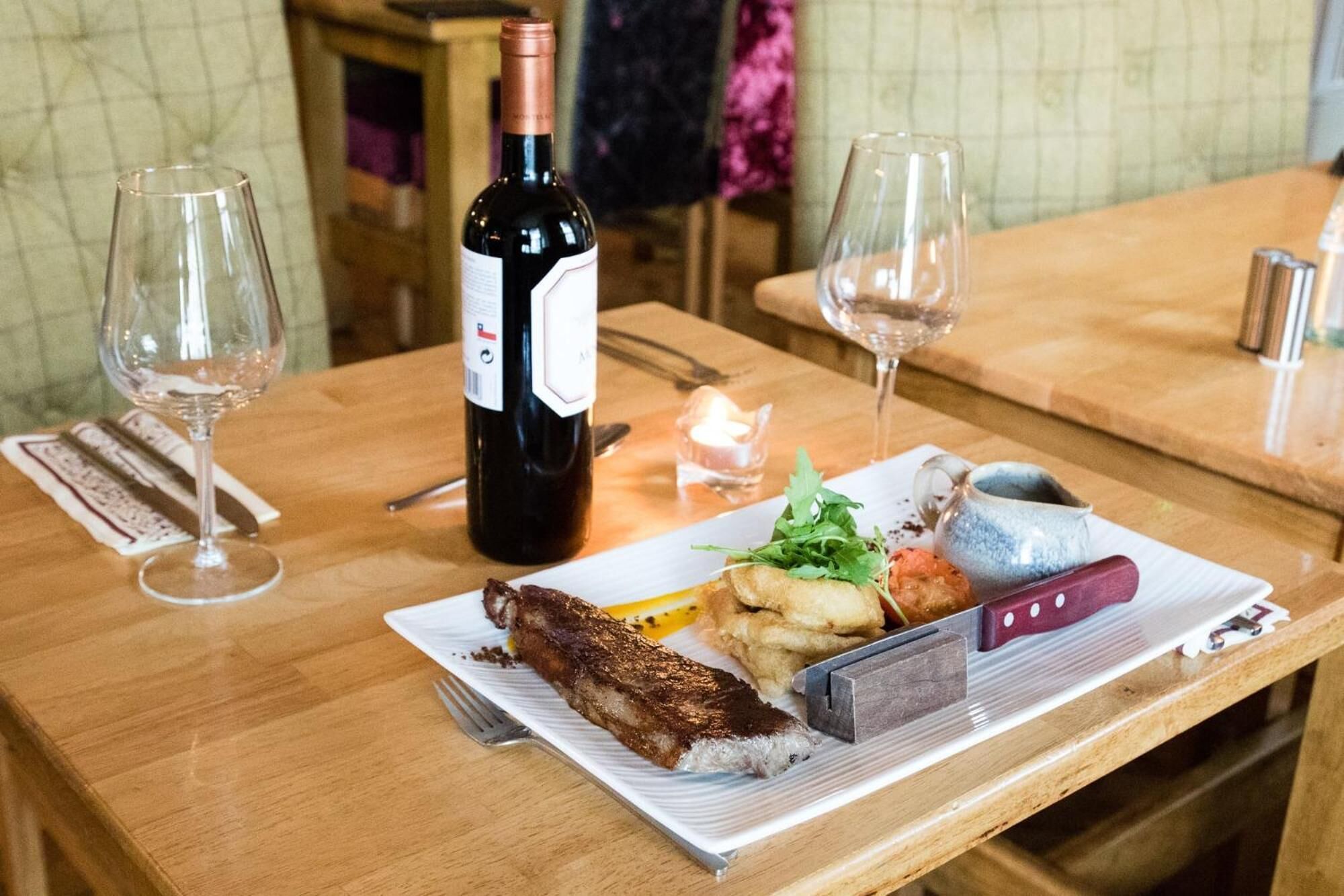 A dining table with a plate of food, a bottle of wine, and two glasses of wine.