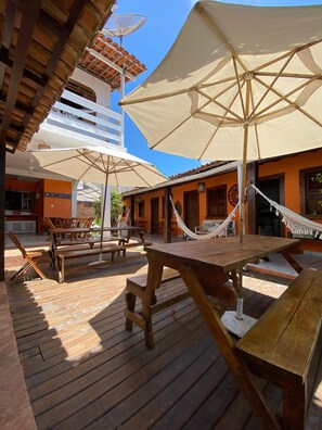 Un patio al aire libre con mesas y bancos de madera, una hamaca y un paraguas grande que da sombra.