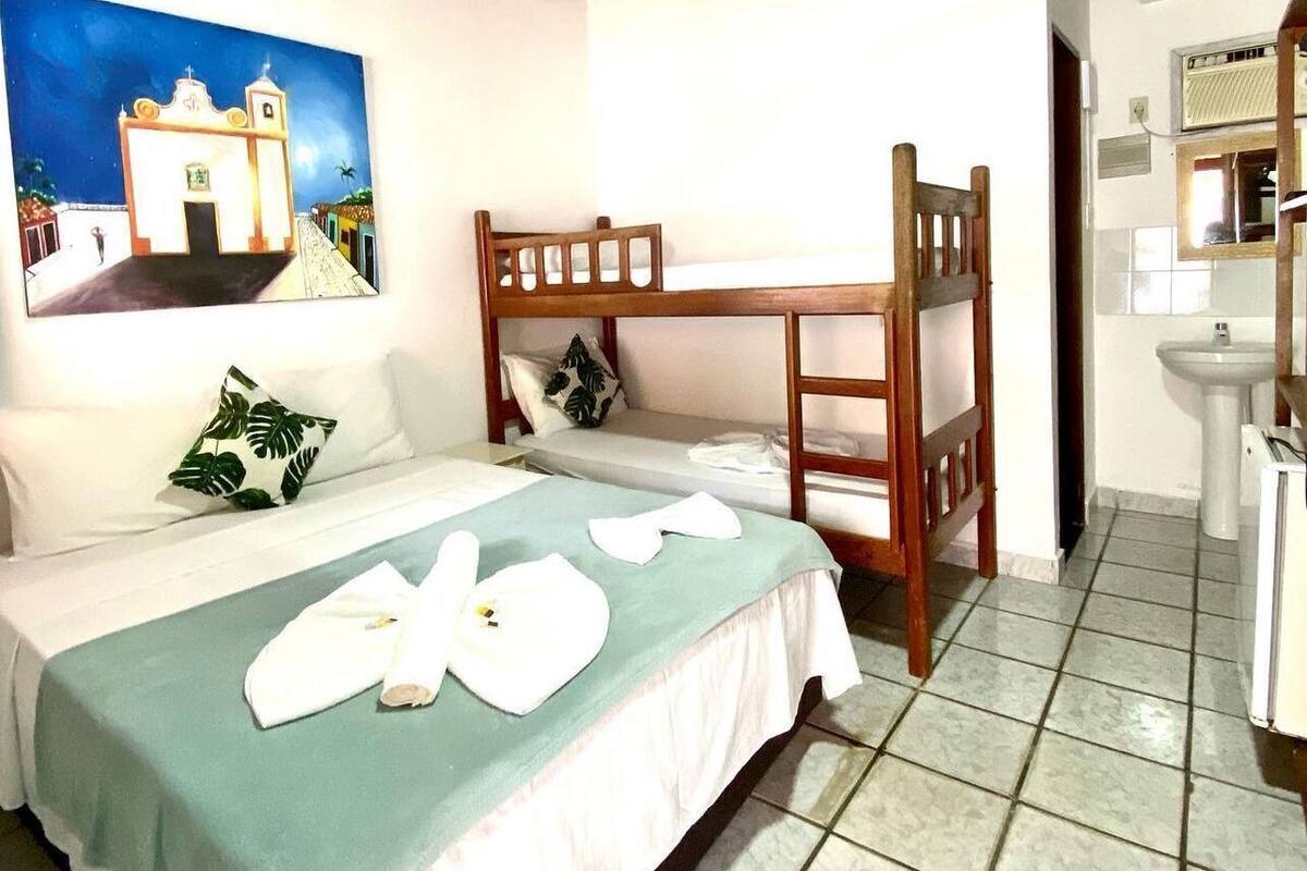 Habitación de hotel con cama, cabecera de madera, ventilador de techo, un cuadro, baño con lavamanos y espejo, y suelo de baldosas.