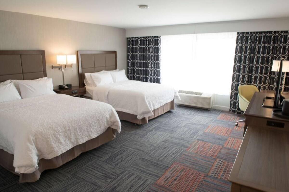 A hotel room with two beds, a desk, a chair, and a large window with patterned curtains.