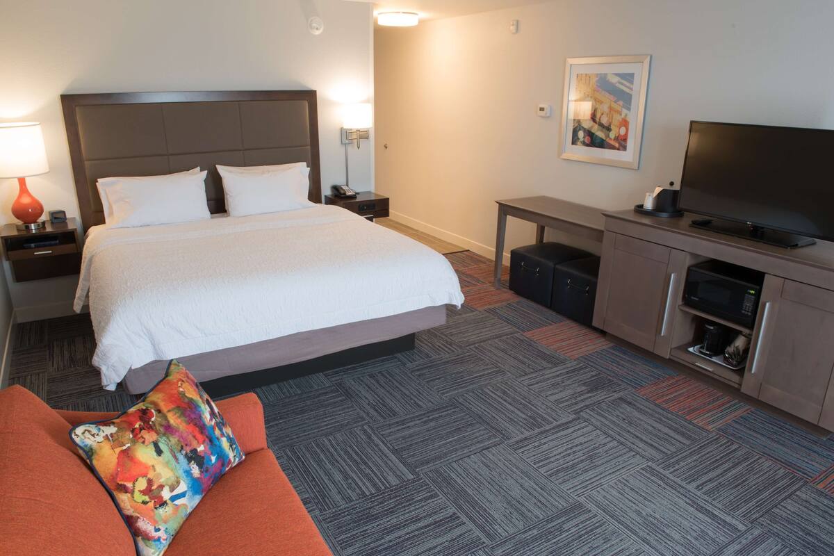 A hotel room with a large bed, a desk with a television, a coffee maker, and a colorful pillow on the couch.