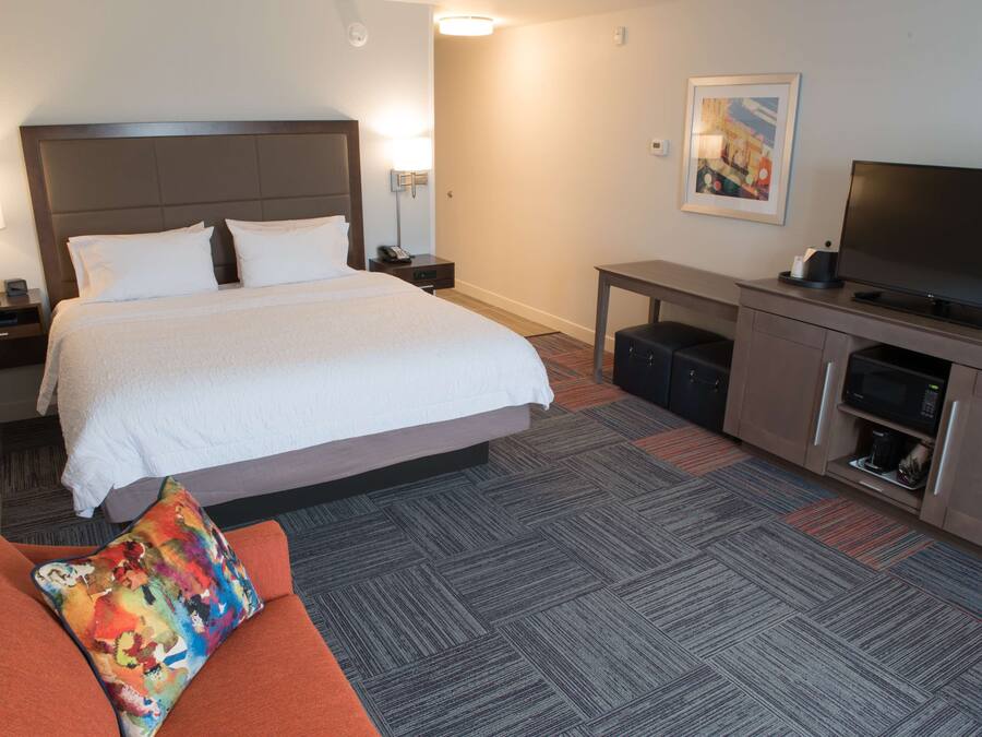 A hotel room with a large bed, a desk with a television, a coffee maker, and a colorful pillow on the couch.