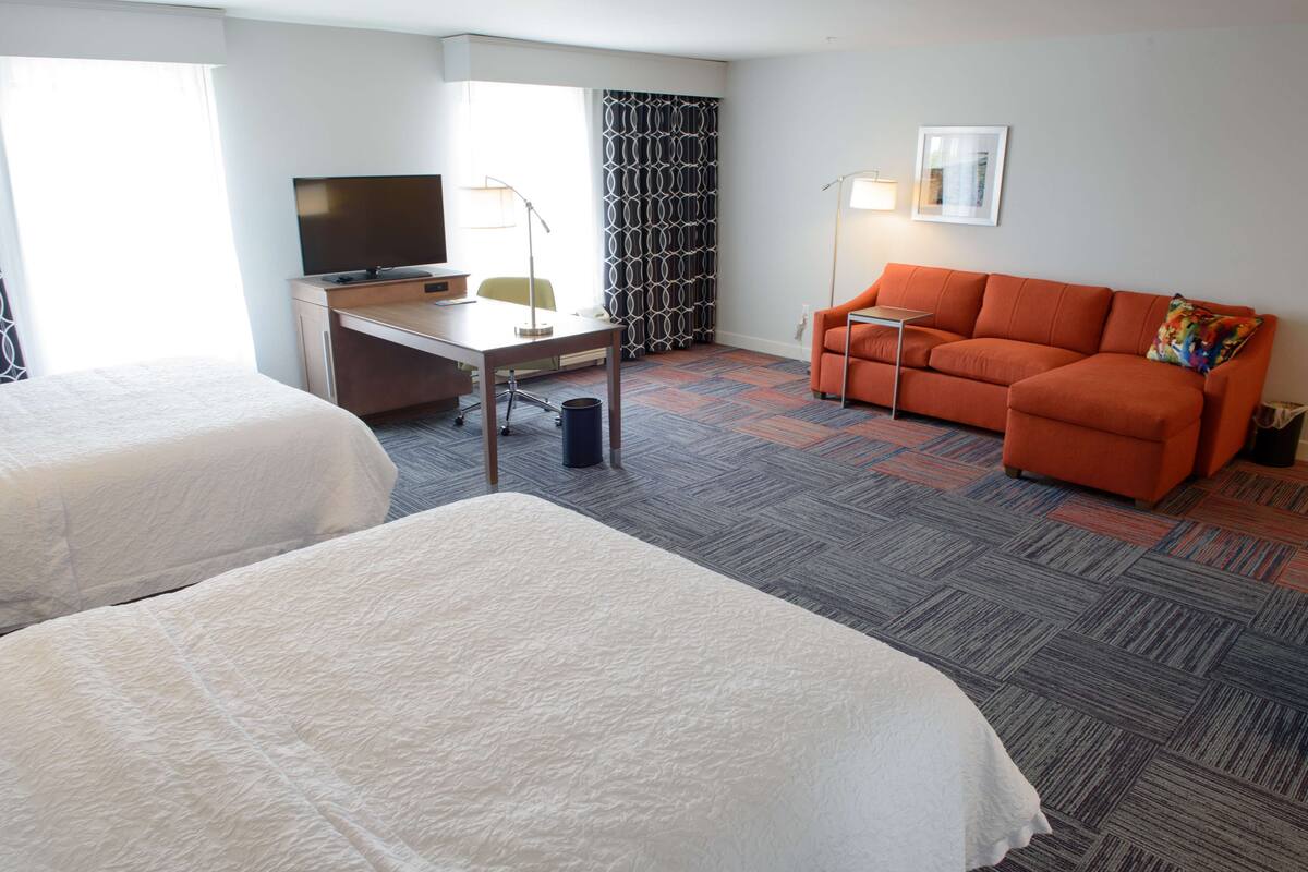 A hotel room with two beds, a desk with a TV, an orange sofa, and a patterned curtain.