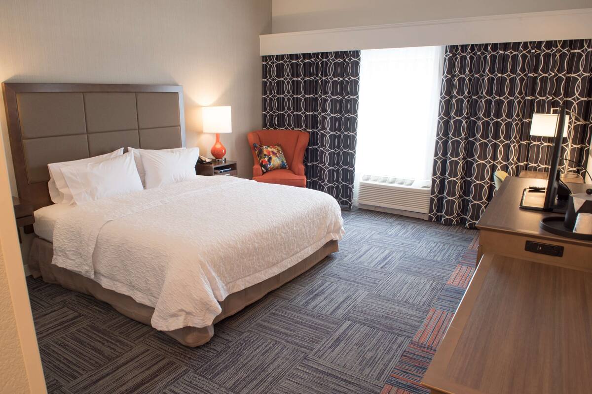 A hotel room with a large bed, a chair, a desk, and a window with patterned curtains.