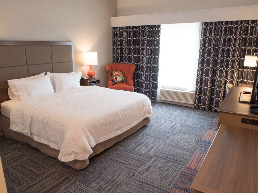A hotel room with a large bed, a chair, a desk, and a window with patterned curtains.