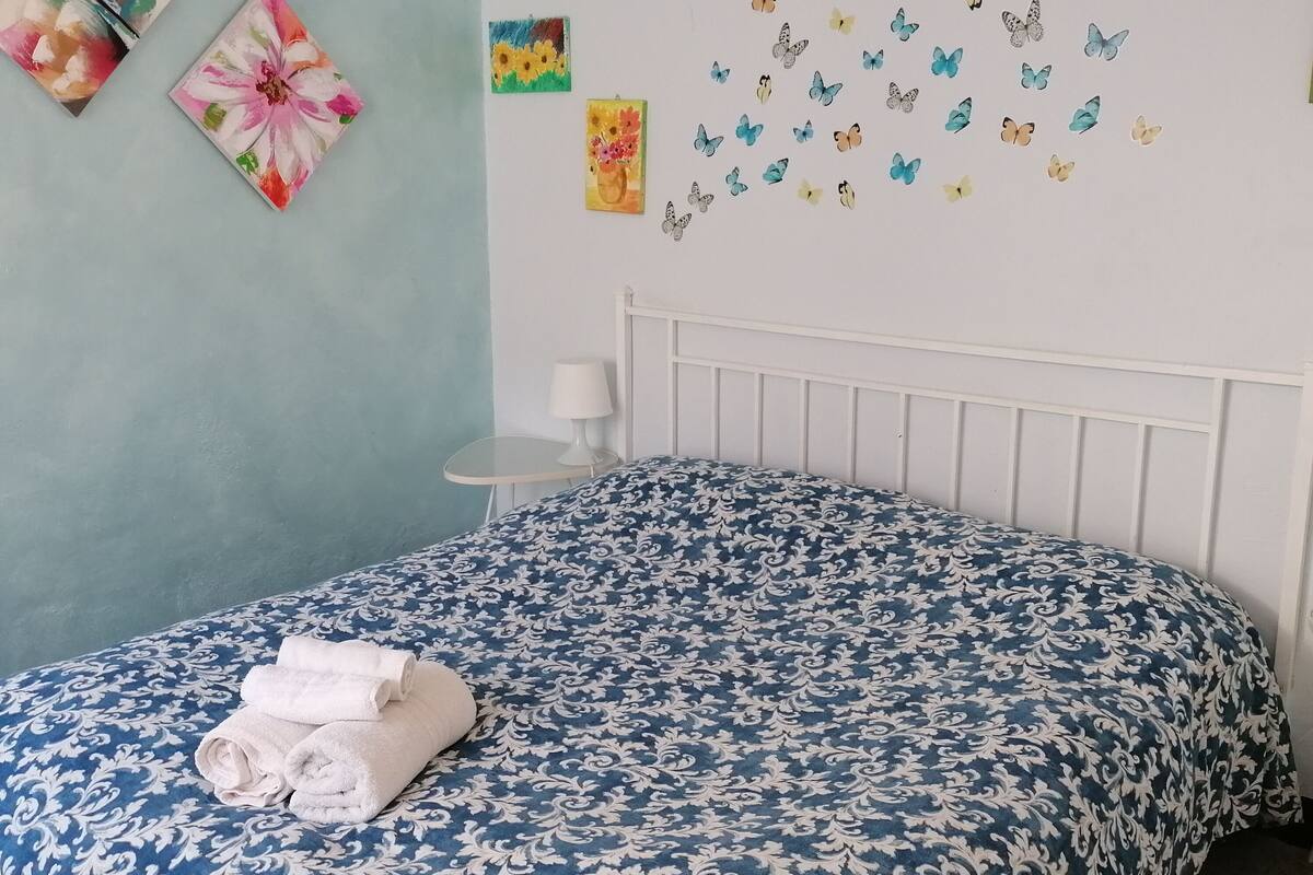 A bedroom with a blue patterned bedspread, two towels on the bed, and wall art including a flower painting and a butterfly decal.