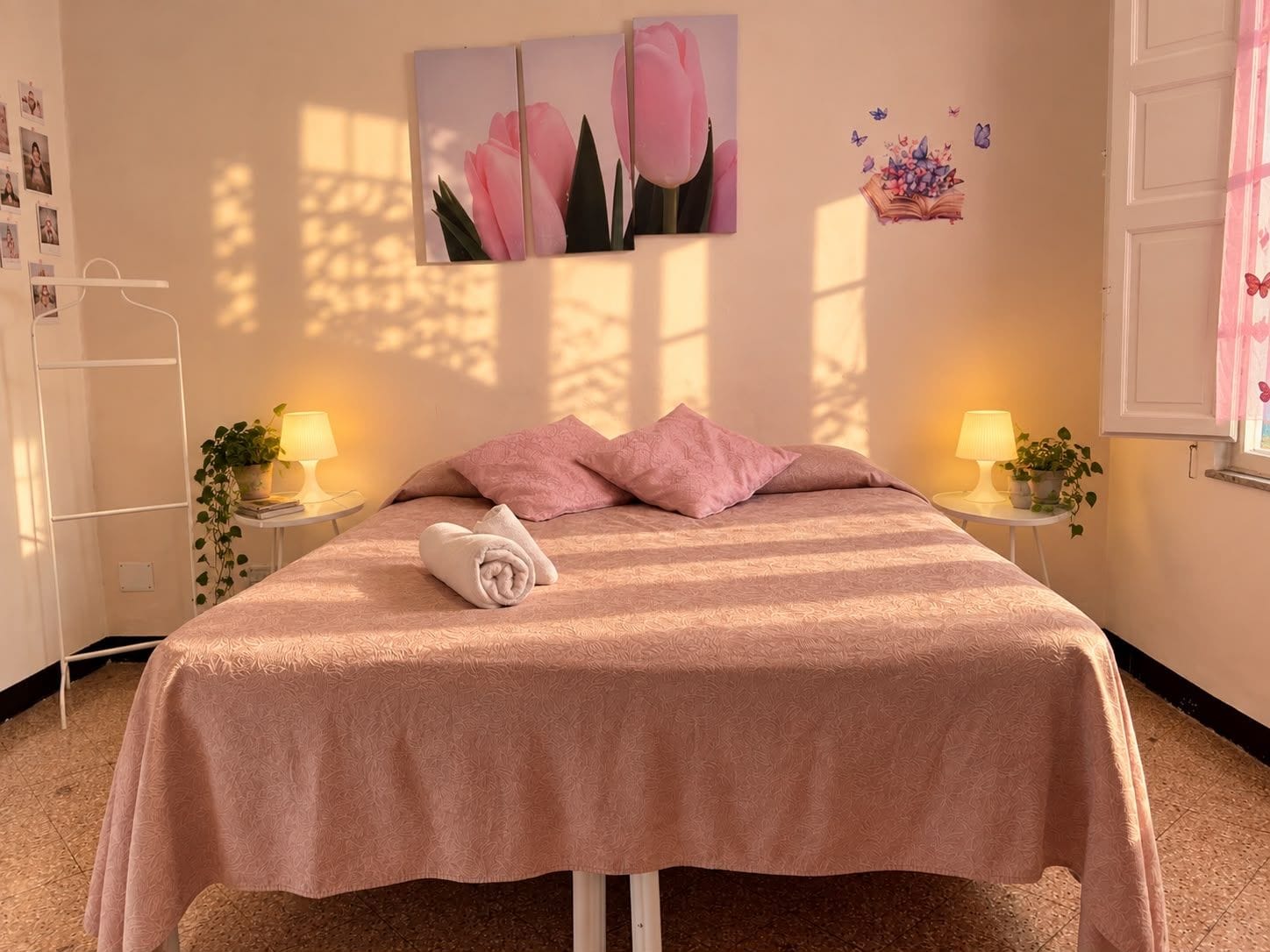 Ein Schlafzimmer mit einem Bett, zwei Nachttischen mit Lampen, einem Fenster mit Vorhängen und Wanddekorationen, darunter ein Bild von Blumen und Schmetterlingen.