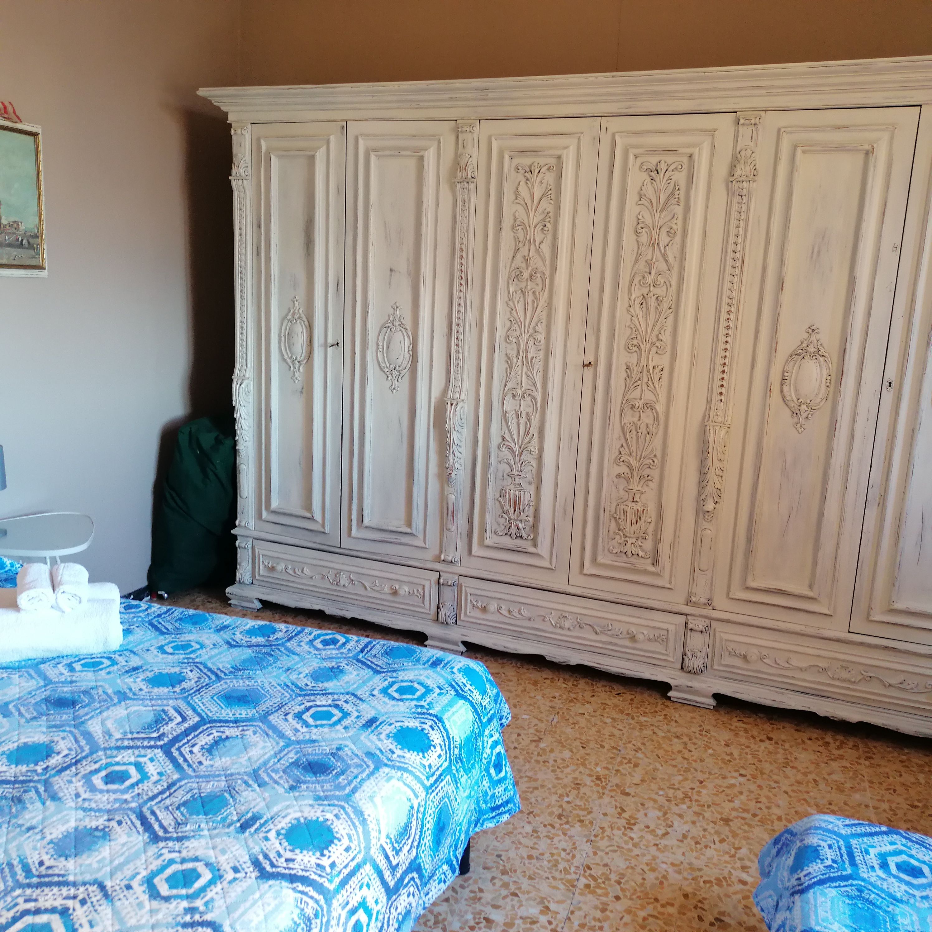 A bedroom with a large white wardrobe, a bed with a patterned blue and white duvet, and a small table with a green bag beside it.