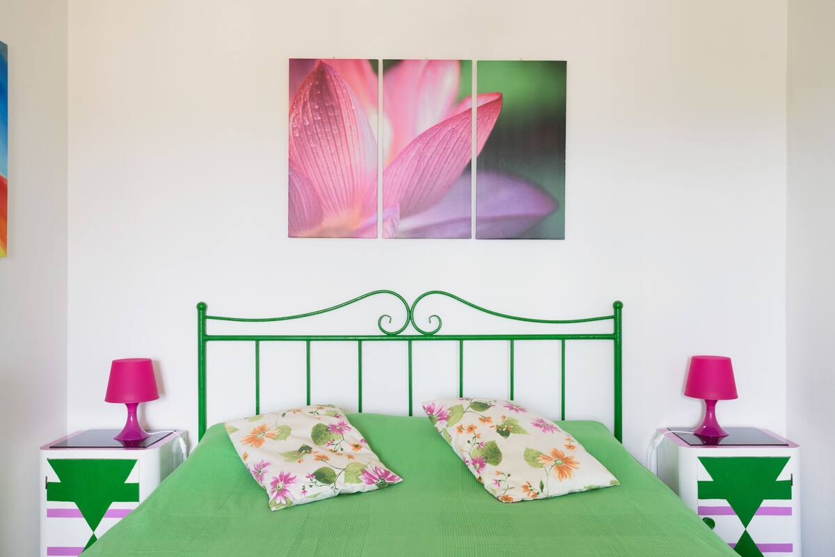 A bedroom with a green bed, floral pillows, a green metal headboard, and pink lamps on matching nightstands.