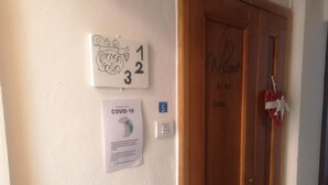 Een gang met een houten deur, een welkomstbord, een bericht over preventie van COVID-19 en een nummerbord.