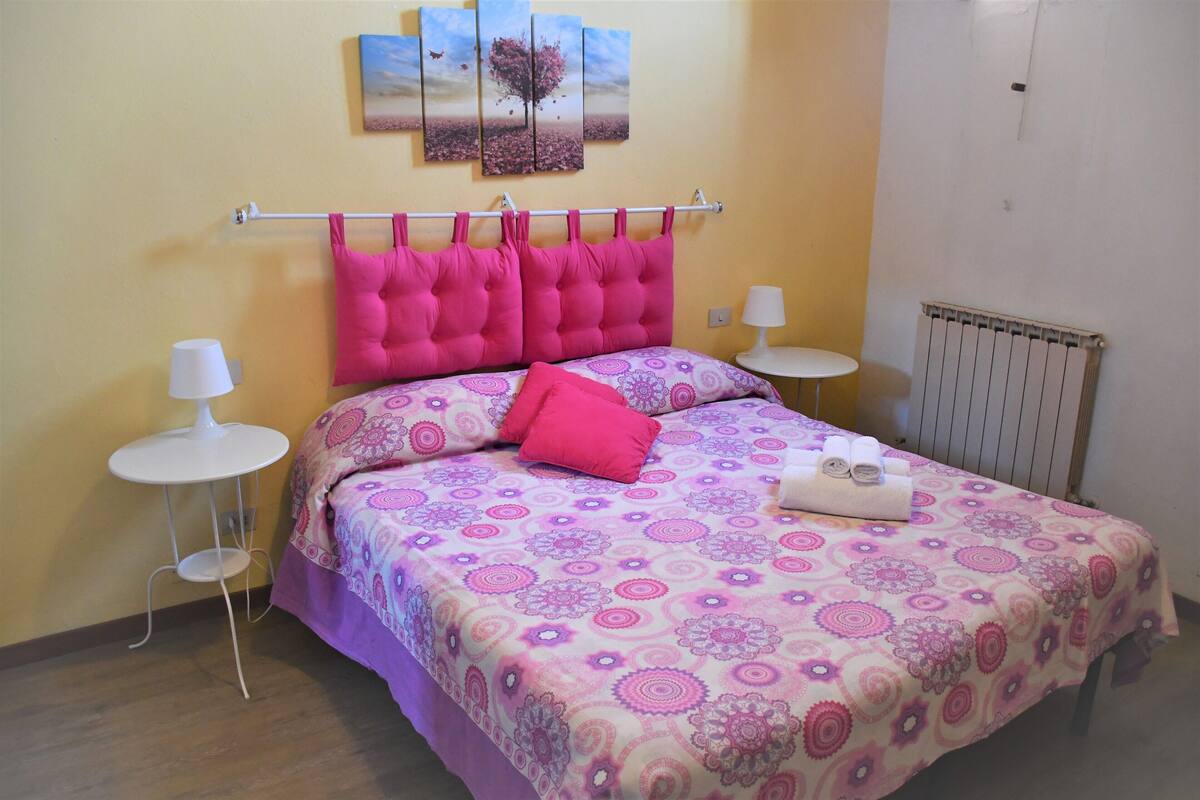 A bedroom with a bed, two bedside tables, a radiator, and a wall with a painting.