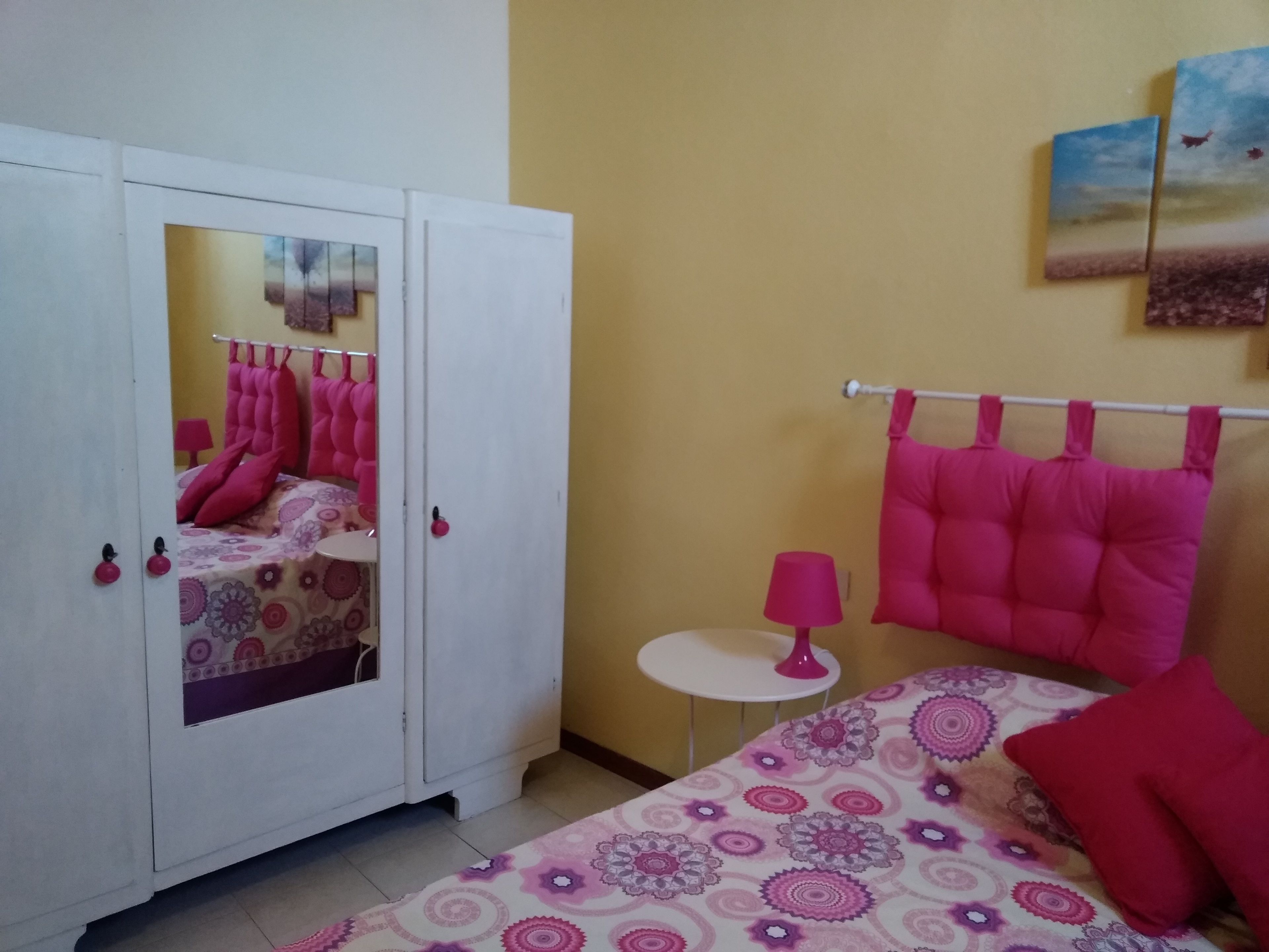 A bedroom with a bed, a wardrobe, a small table, and a wall-mounted lamp.