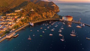 An aerial view of a coastal town with a marina filled with boats, a prominent building with a red roof, and a hilly landscape in the background. - Seaport Village Inn Avalon (Avalon)